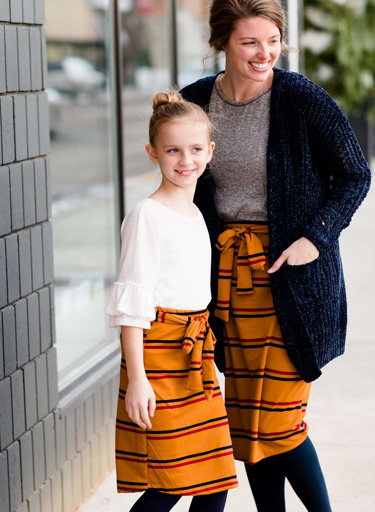 Woman wearing a modest mustard and burgandy striped midi skirt. This skirt has a fabric belt that is removable and is also mommy and me styled. This skirt is paired with tights and boots.