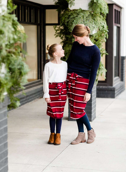 Young girl wearing a burgandy and mustard striped midi skirt with removable belt. This is also a mommy and me skirt. It is paired with a white ruffle top, leggings and brown boots.