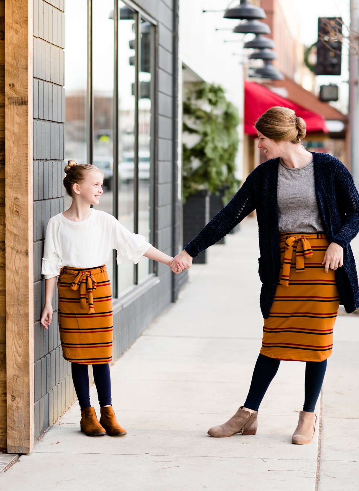 Young girl wearing a burgandy and mustard striped midi skirt with removable belt. This is also a mommy and me skirt. It is paired with a white ruffle top, leggings and brown boots.