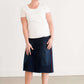 Woman wearing a dark indigo wash below the knee skirt that has red piping along the sides. She also is wearing a white tee shirt and tan sneakers.