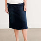 Woman wearing a dark indigo wash below the knee skirt that has red piping along the sides. She also is wearing a white tee shirt and tan sneakers.