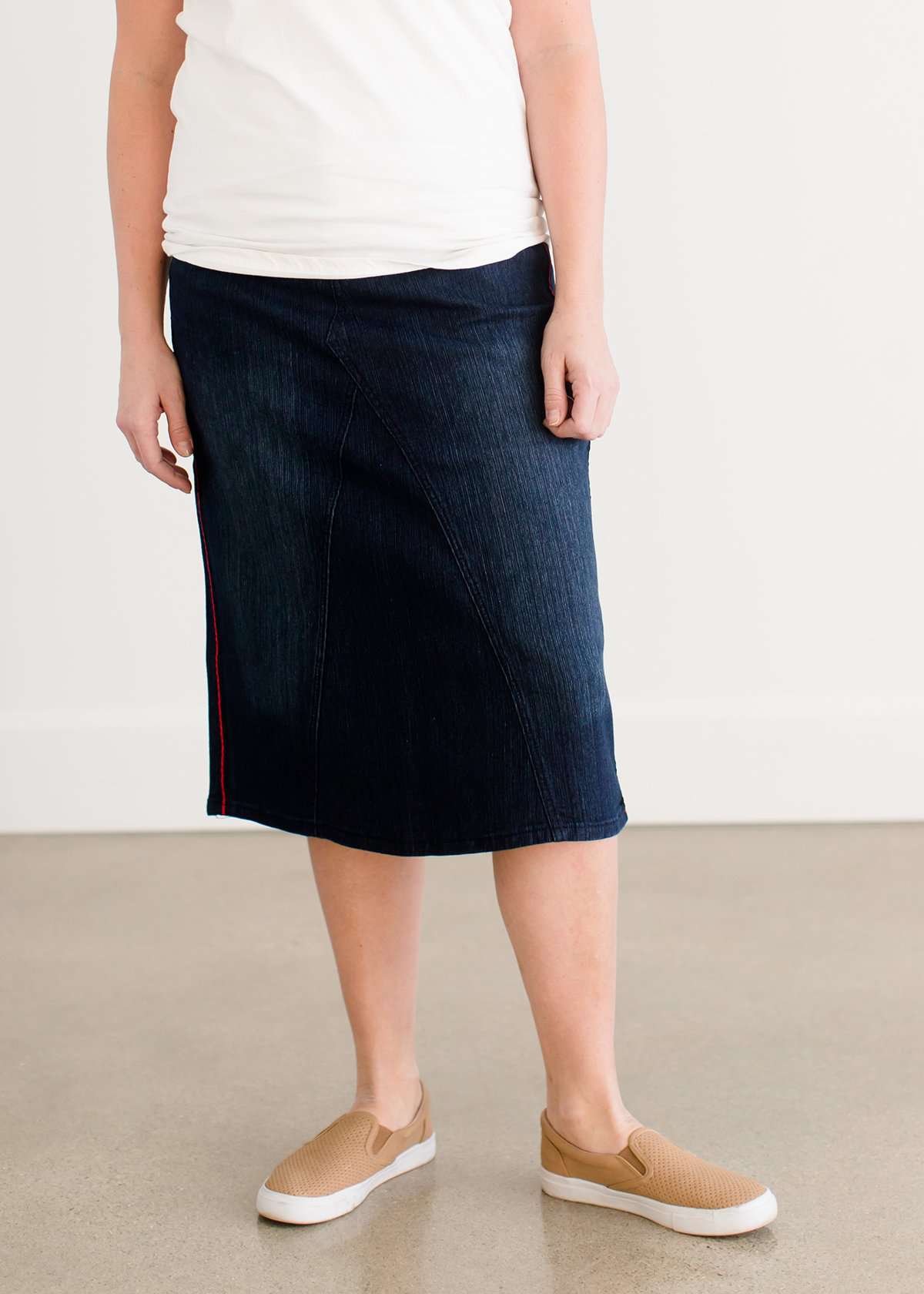 Woman wearing a dark indigo wash below the knee skirt that has red piping along the sides. She also is wearing a white tee shirt and tan sneakers.