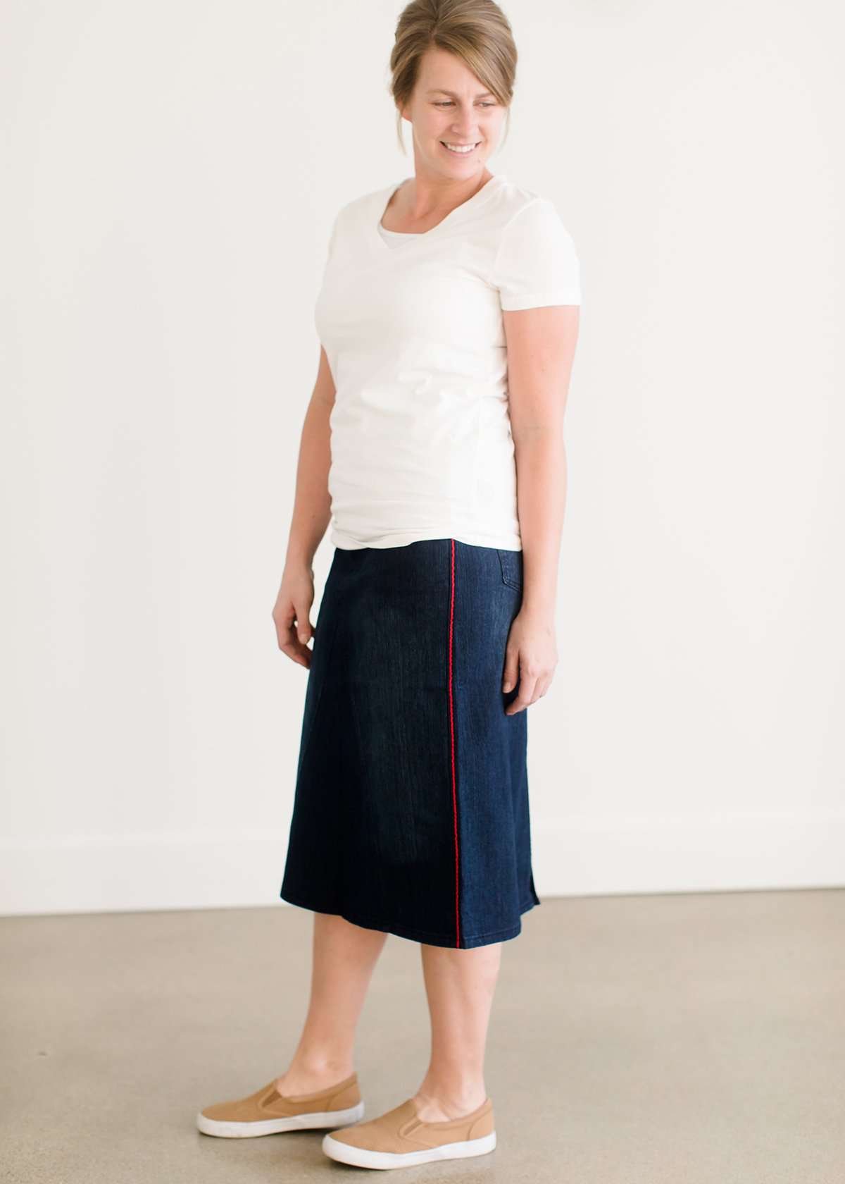 Woman wearing a dark indigo wash below the knee skirt that has red piping along the sides. She also is wearing a white tee shirt and tan sneakers.