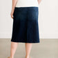 Woman wearing a dark indigo wash below the knee skirt that has red piping along the sides. She also is wearing a white tee shirt and tan sneakers.