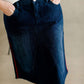 Woman wearing a dark indigo wash below the knee skirt that has red piping along the sides. She also is wearing a white tee shirt and tan sneakers.