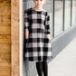 Young girl wearing a black and white buffalo check midi dress. This dress has 3/4 sleeves and front pockets. It is paired with black leggings and black booties.