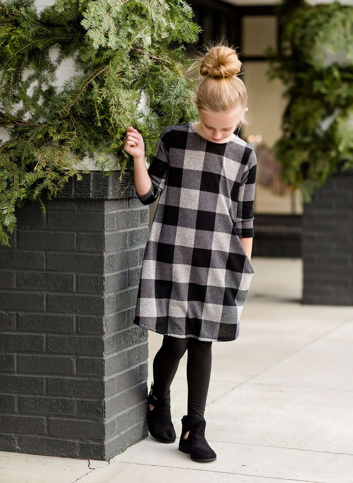 Young girl wearing a black and white buffalo check midi dress. This dress has 3/4 sleeves and front pockets. It is paired with black leggings and black booties.