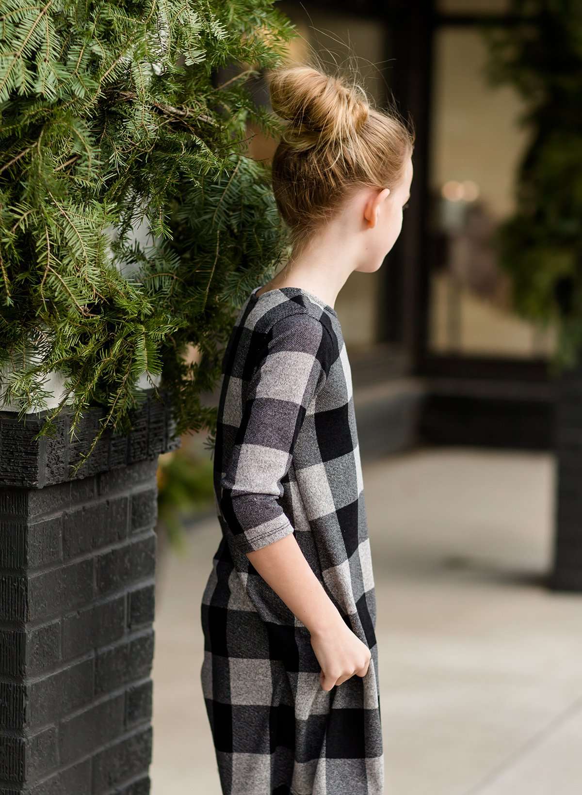 Young girl wearing a black and white buffalo check midi dress. This dress has 3/4 sleeves and front pockets. It is paired with black leggings and black booties.
