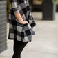 Young girl wearing a black and white buffalo check midi dress. This dress has 3/4 sleeves and front pockets. It is paired with black leggings and black booties.