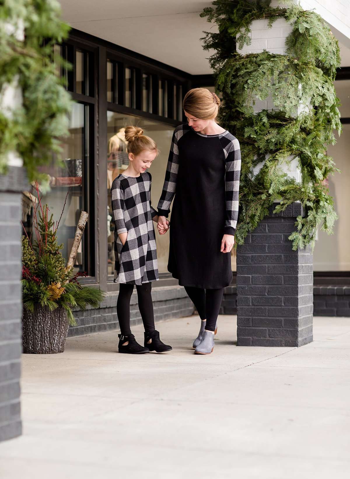 Young girl wearing a black and white buffalo check midi dress. This dress has 3/4 sleeves and front pockets. It is paired with black leggings and black booties.