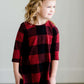 Young girl wearing a black and red buffalo check midi dress. This dress has 3/4 sleeves and front pockets. It is paired with black leggings and black booties.