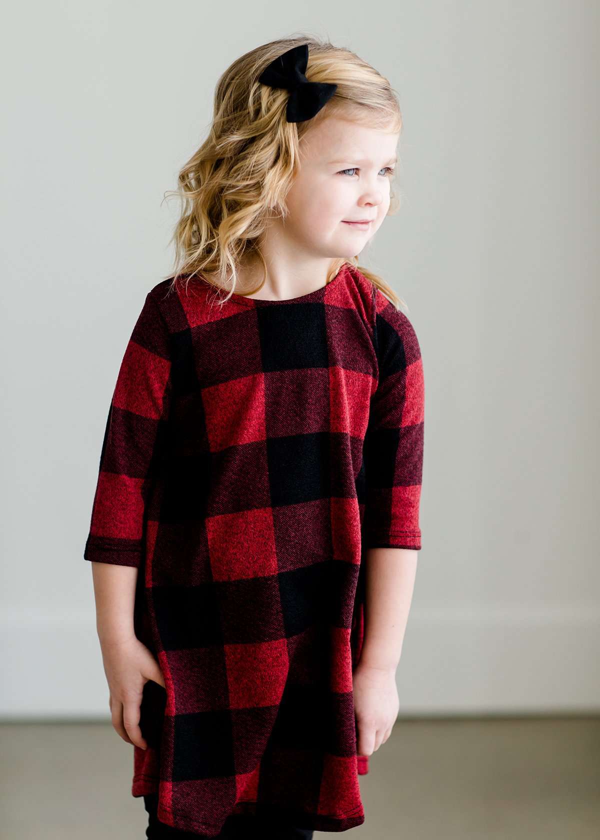 Young girl wearing a black and red buffalo check midi dress. This dress has 3/4 sleeves and front pockets. It is paired with black leggings and black booties.