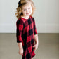 Young girl wearing a black and red buffalo check midi dress. This dress has 3/4 sleeves and front pockets. It is paired with black leggings and black booties.