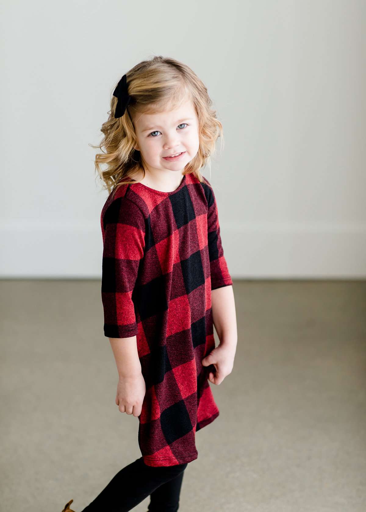 Young girl wearing a black and red buffalo check midi dress. This dress has 3/4 sleeves and front pockets. It is paired with black leggings and black booties.