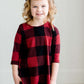 Young girl wearing a black and red buffalo check midi dress. This dress has 3/4 sleeves and front pockets. It is paired with black leggings and black booties.
