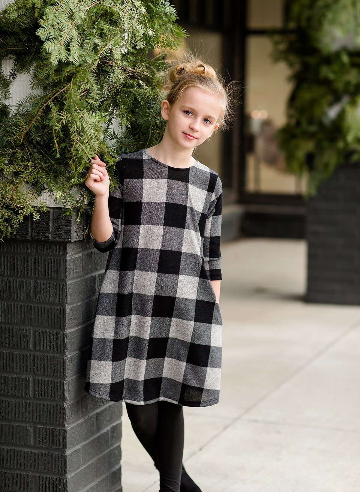 Young girl wearing a black and white buffalo check midi dress. This dress has 3/4 sleeves and front pockets. It is paired with black leggings and black booties.