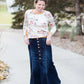 Woman wearing a long denim skirt without a slit that has buttons up the front and is a flare, a-line style.