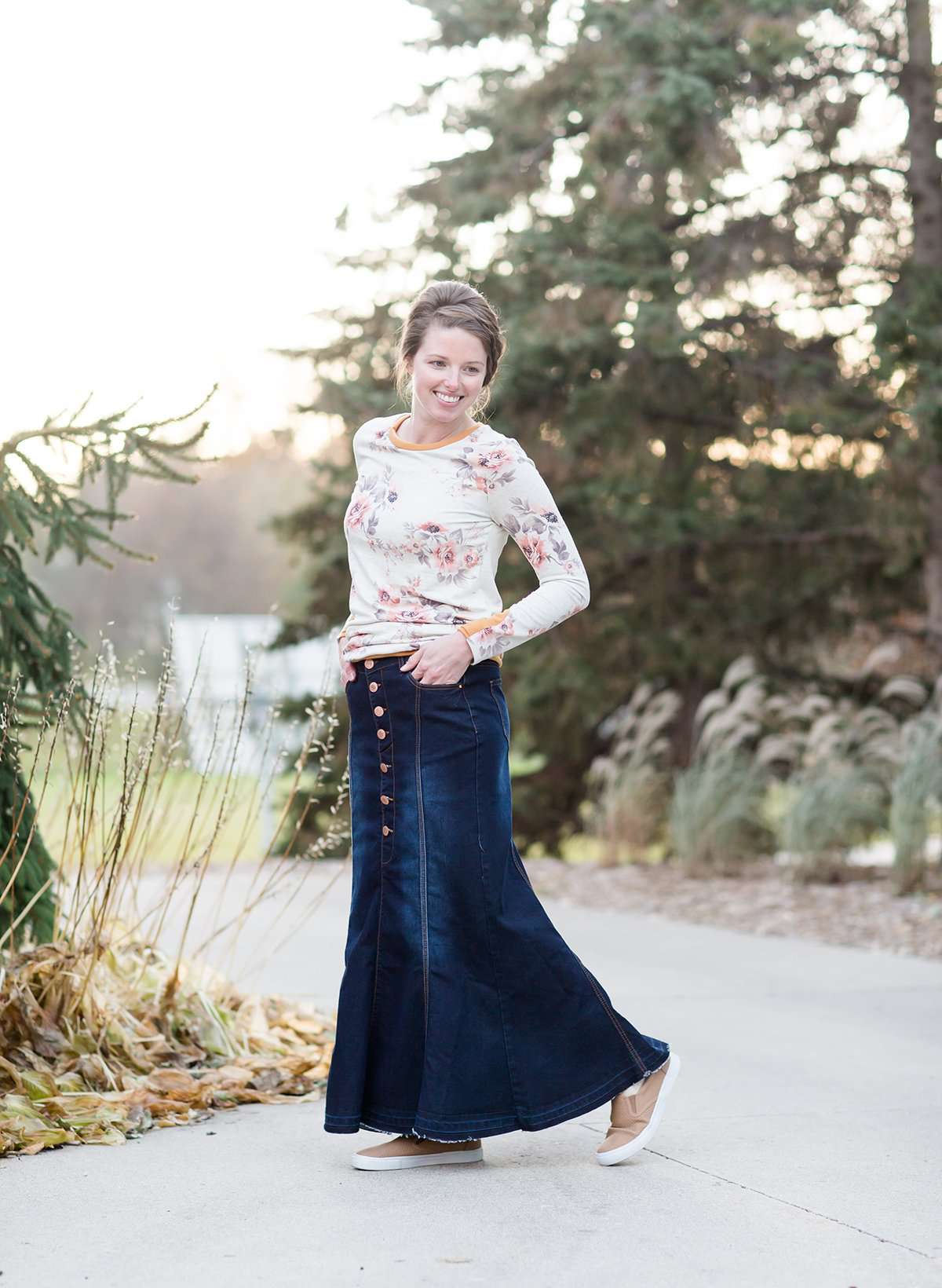 Woman wearing a long denim skirt without a slit that has buttons up the front and is a flare, a-line style.