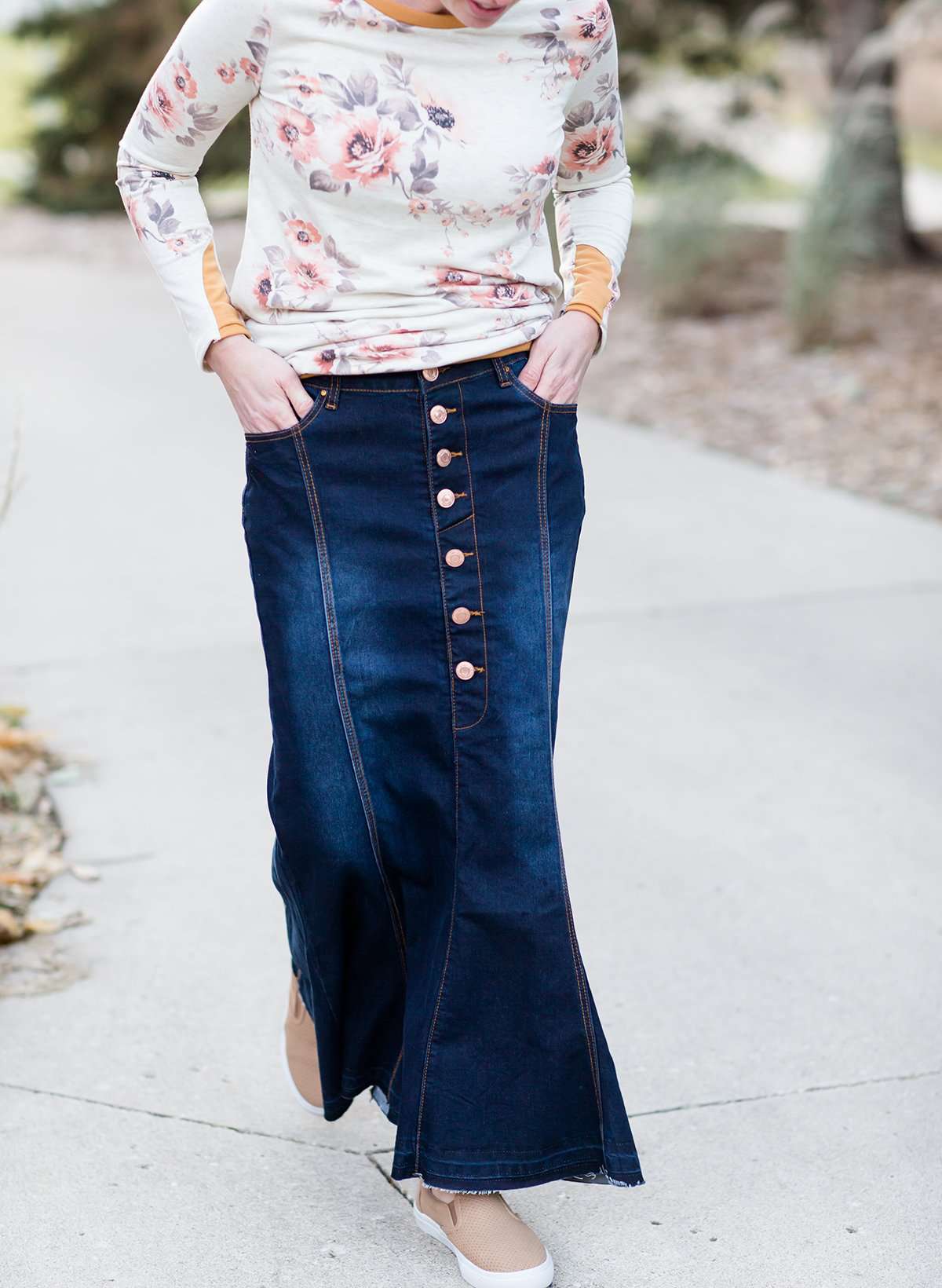 Woman wearing a long denim skirt without a slit that has buttons up the front and is a flare, a-line style.