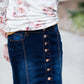 Woman wearing a long denim skirt without a slit that has buttons up the front and is a flare, a-line style.