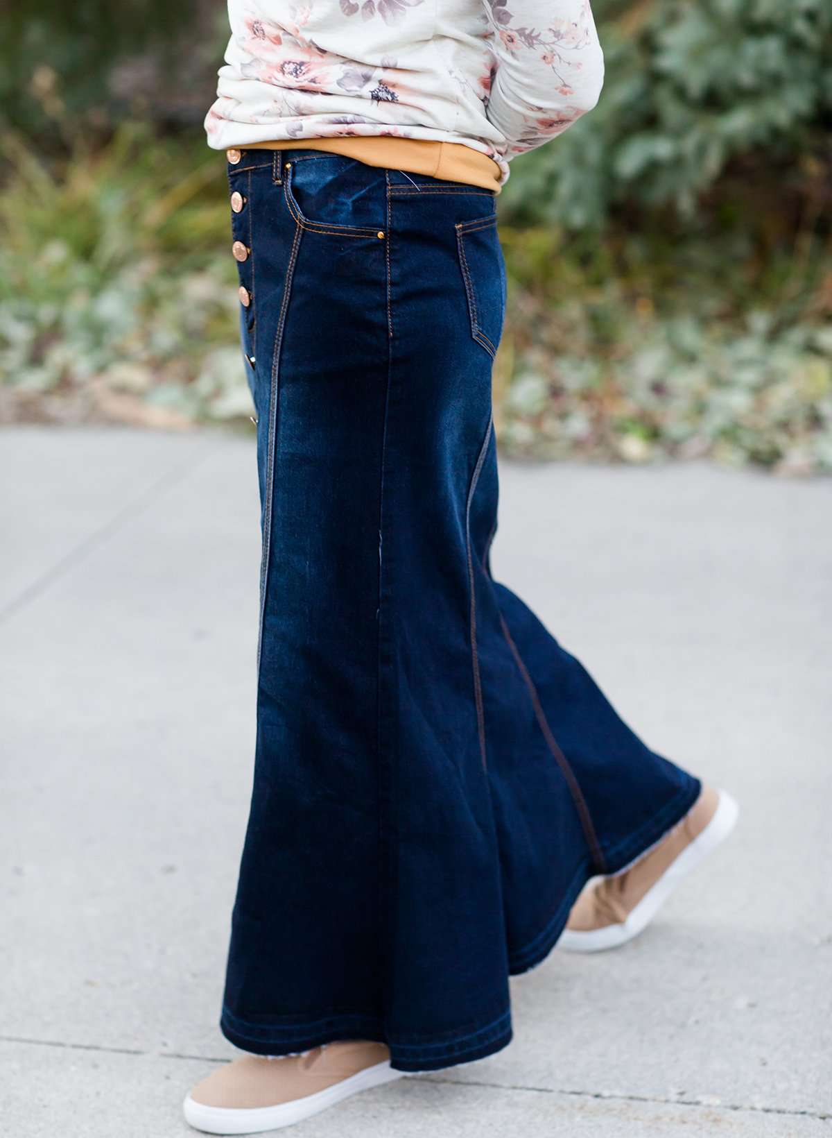 Woman wearing a long denim skirt without a slit that has buttons up the front and is a flare, a-line style.