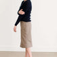 Woman wearing a khaki midi length skirt with faux buttons running down the front. It is paired with a navy sweater and copper, suede slide shoes.