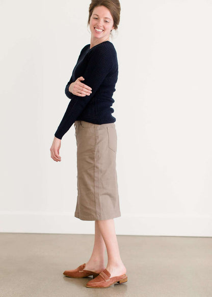 Woman wearing a khaki midi length skirt with faux buttons running down the front. It is paired with a navy sweater and copper, suede slide shoes.
