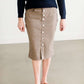 Woman wearing a khaki midi length skirt with faux buttons running down the front. It is paired with a navy sweater and copper, suede slide shoes.
