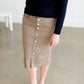 Woman wearing a khaki midi length skirt with faux buttons running down the front. It is paired with a navy sweater and copper, suede slide shoes.