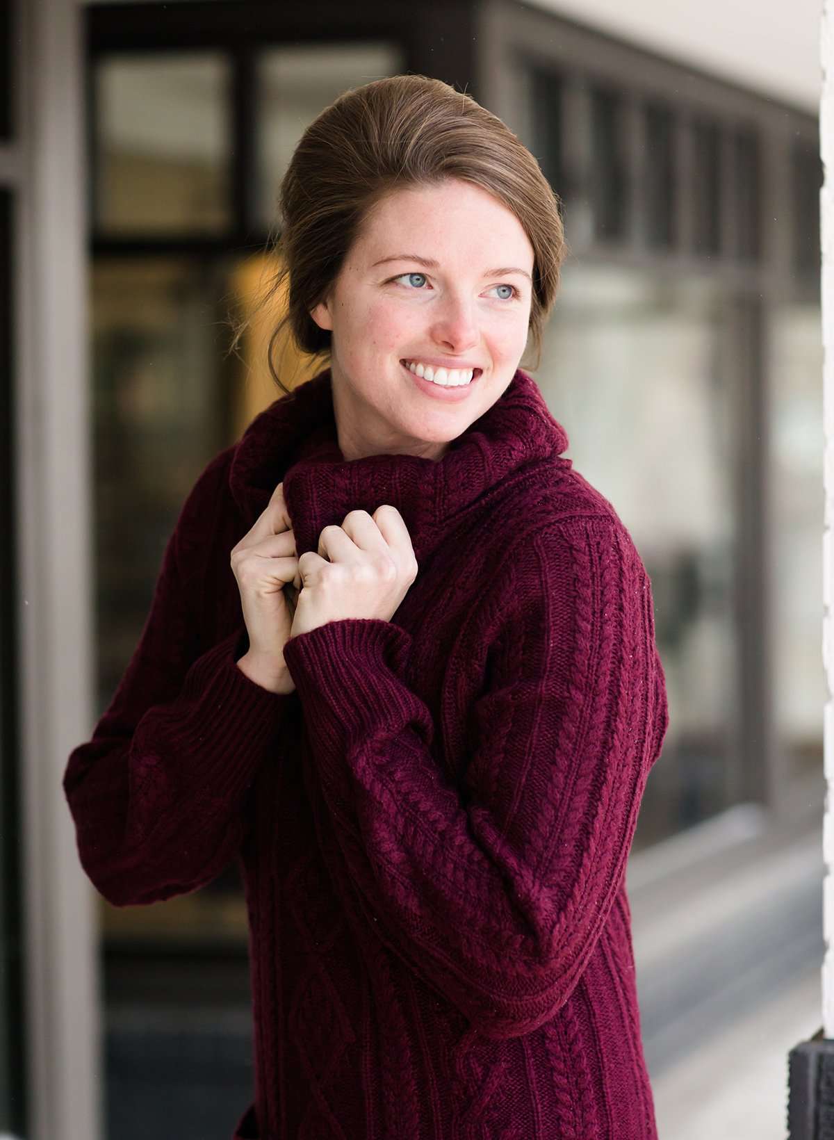 Woman wearing a cableknit, cowlneck sweater dress in taupe or burgandy. This cozy dress is below the knees and paired with tall, suede riding boots.