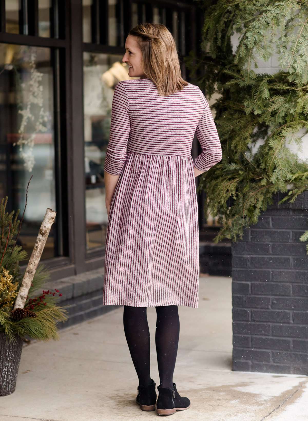 Woman wearing a wine and gray striped modest midi dress with front, square pockets. She also has black leggings and sole society black boots on.