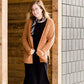 Woman wearing an open front cardigan that is chenille and camel in color. She is also wearing it over a black dress and brown riding boots. She is standing on the stairs at Inherit Clothing Company.
