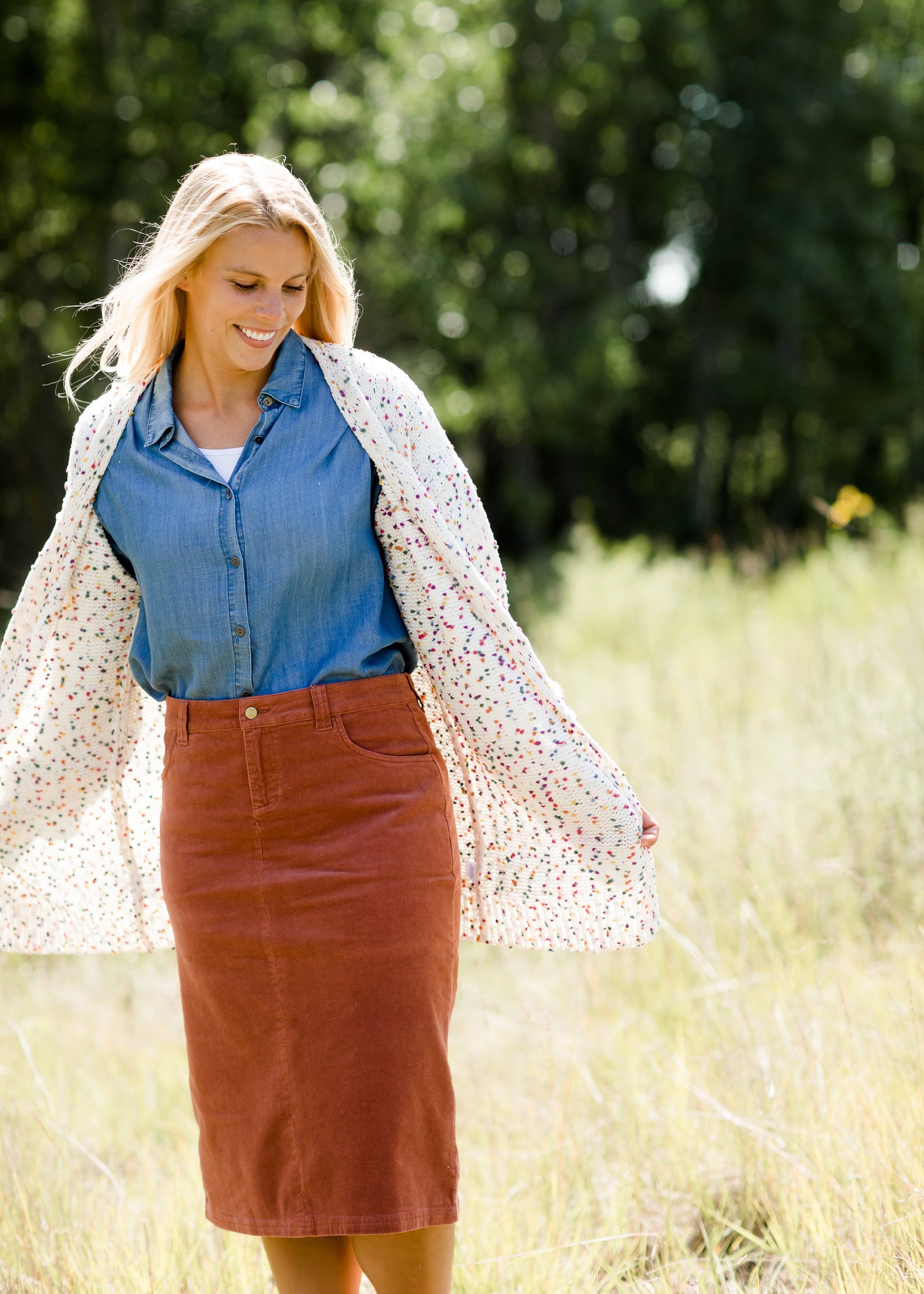 Woman wearing a below the knee corduroy skirt. This skirt is a fin corduroy with a gold button snap, no slit and no back pockets. It comes in burgundy, teal, burnt orange and dark khaki.