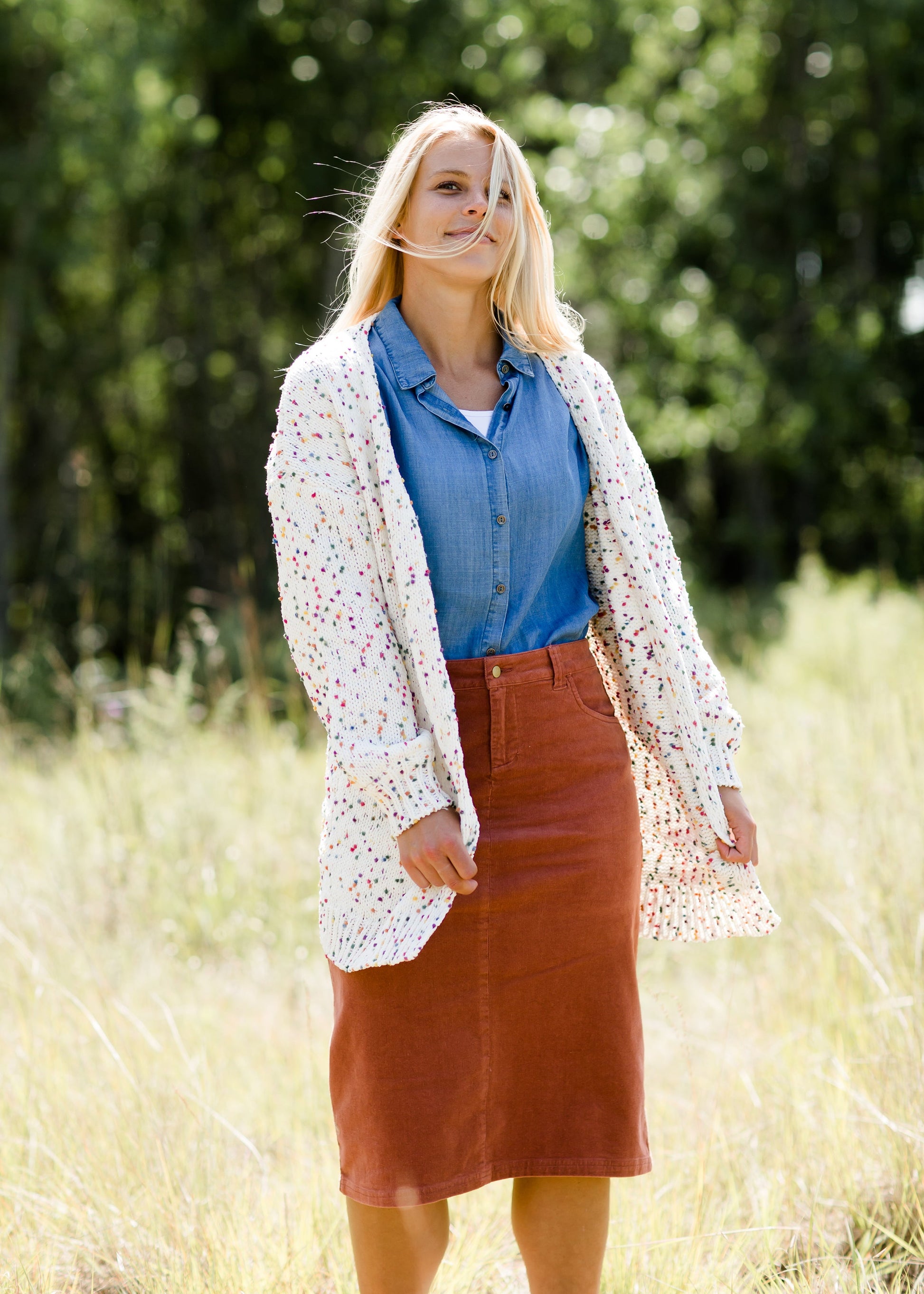 Woman wearing a below the knee corduroy skirt. This skirt is a fin corduroy with a gold button snap, no slit and no back pockets. It comes in burgundy, teal, burnt orange and dark khaki.