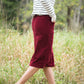 Woman wearing a below the knee corduroy skirt. This skirt is a fin corduroy with a gold button snap, no slit and no back pockets. It comes in burgundy, teal, burnt orange and dark khaki.