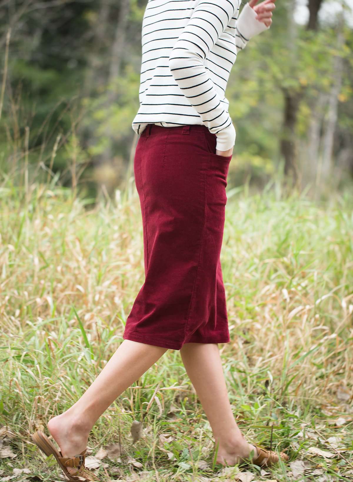 Woman wearing a below the knee corduroy skirt. This skirt is a fin corduroy with a gold button snap, no slit and no back pockets. It comes in burgundy, teal, burnt orange and dark khaki.