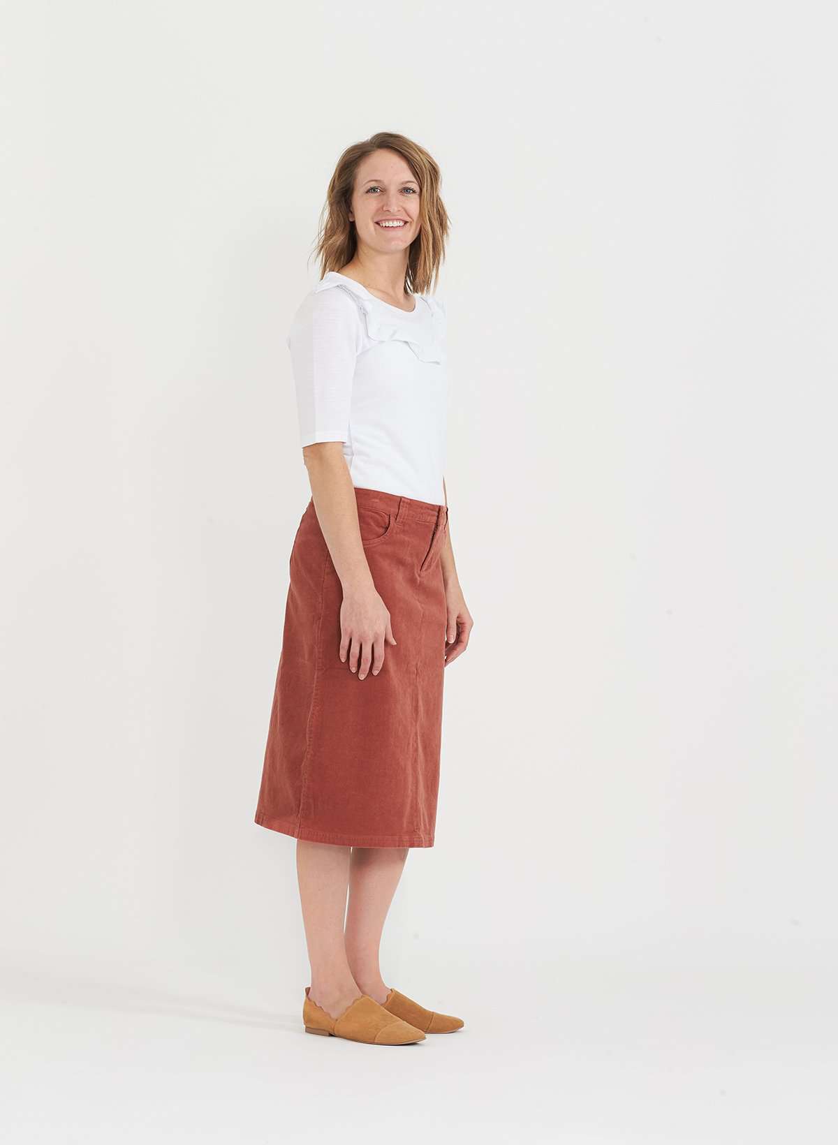 Woman wearing a below the knee corduroy skirt. This skirt is a fin corduroy with a gold button snap, no slit and no back pockets. It comes in burgundy, teal, burnt orange and dark khaki.