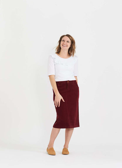 Woman wearing a below the knee corduroy skirt. This skirt is a fin corduroy with a gold button snap, no slit and no back pockets. It comes in burgundy, teal, burnt orange and dark khaki.