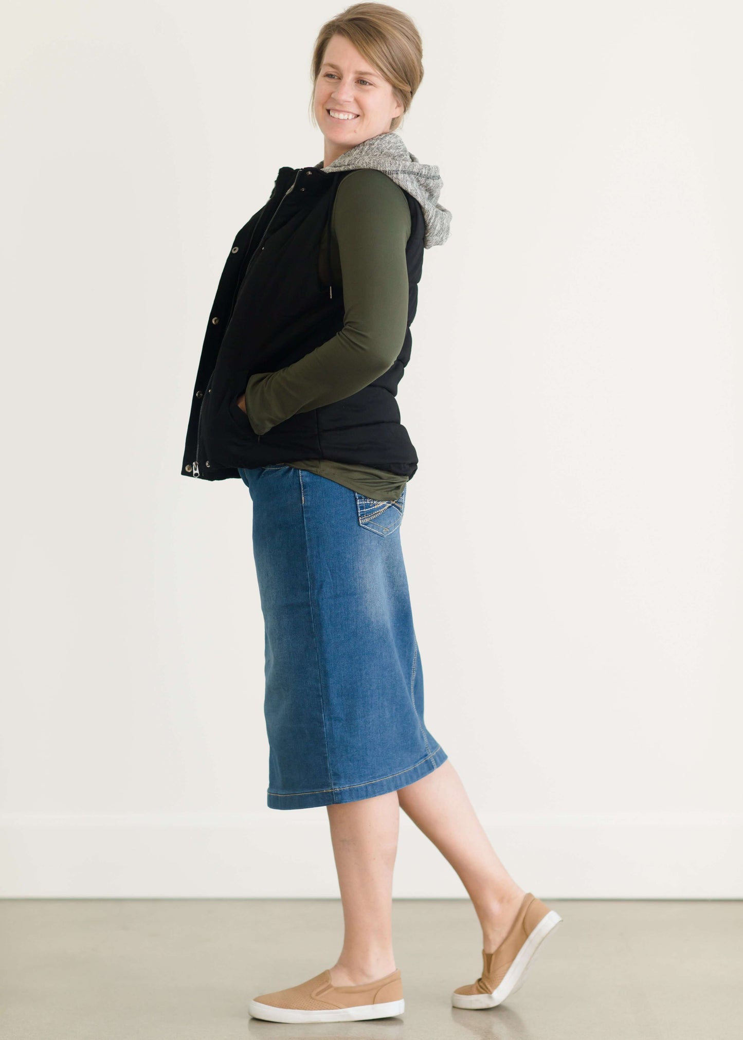 Woman wearing a mid length jean skirt that is a pencil fit. It has a jewel on the back pockets and is paired with sneakers and a olive long sleeve tee and black vest.
