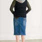 Woman wearing a mid length jean skirt that is a pencil fit. It has a jewel on the back pockets and is paired with sneakers and a olive long sleeve tee and black vest.