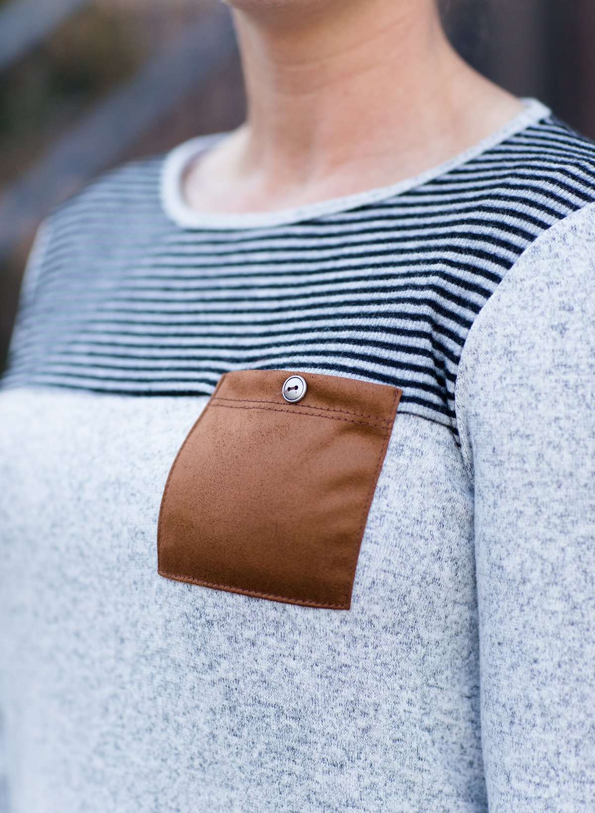 Woman wearing a gray color block stripe top with a brown, suede pocket with a button feature.