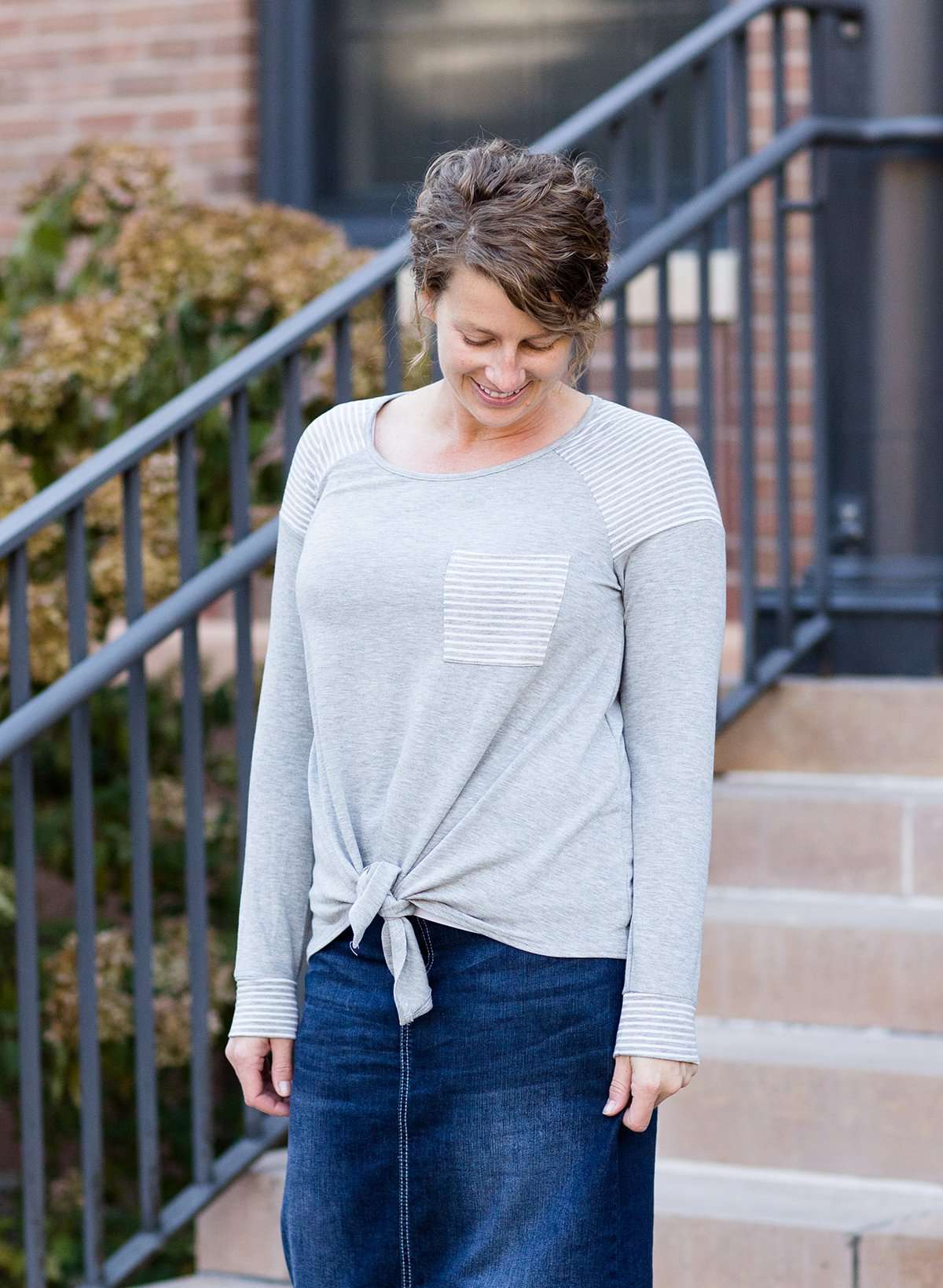 Woman wearing a gray tri blend top that has a striped pocket and a front tie.