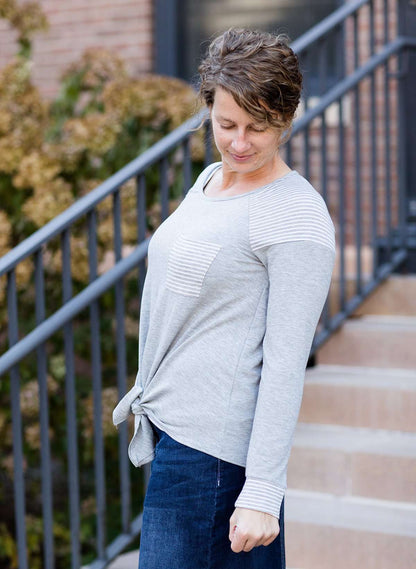 Woman wearing a gray tri blend top that has a striped pocket and a front tie.