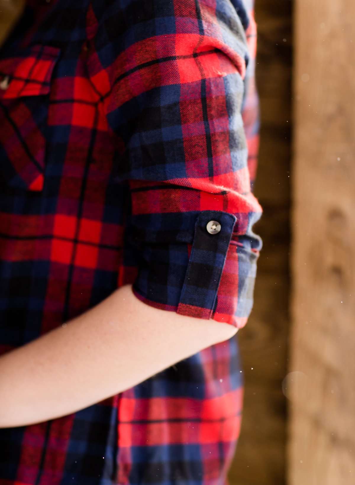 Woman wearing a cozy, checkered plaid top. This button up plaid shirt features roll tab sleeves and is black, navy and red. It is also paired with a below the knee jean skirt.