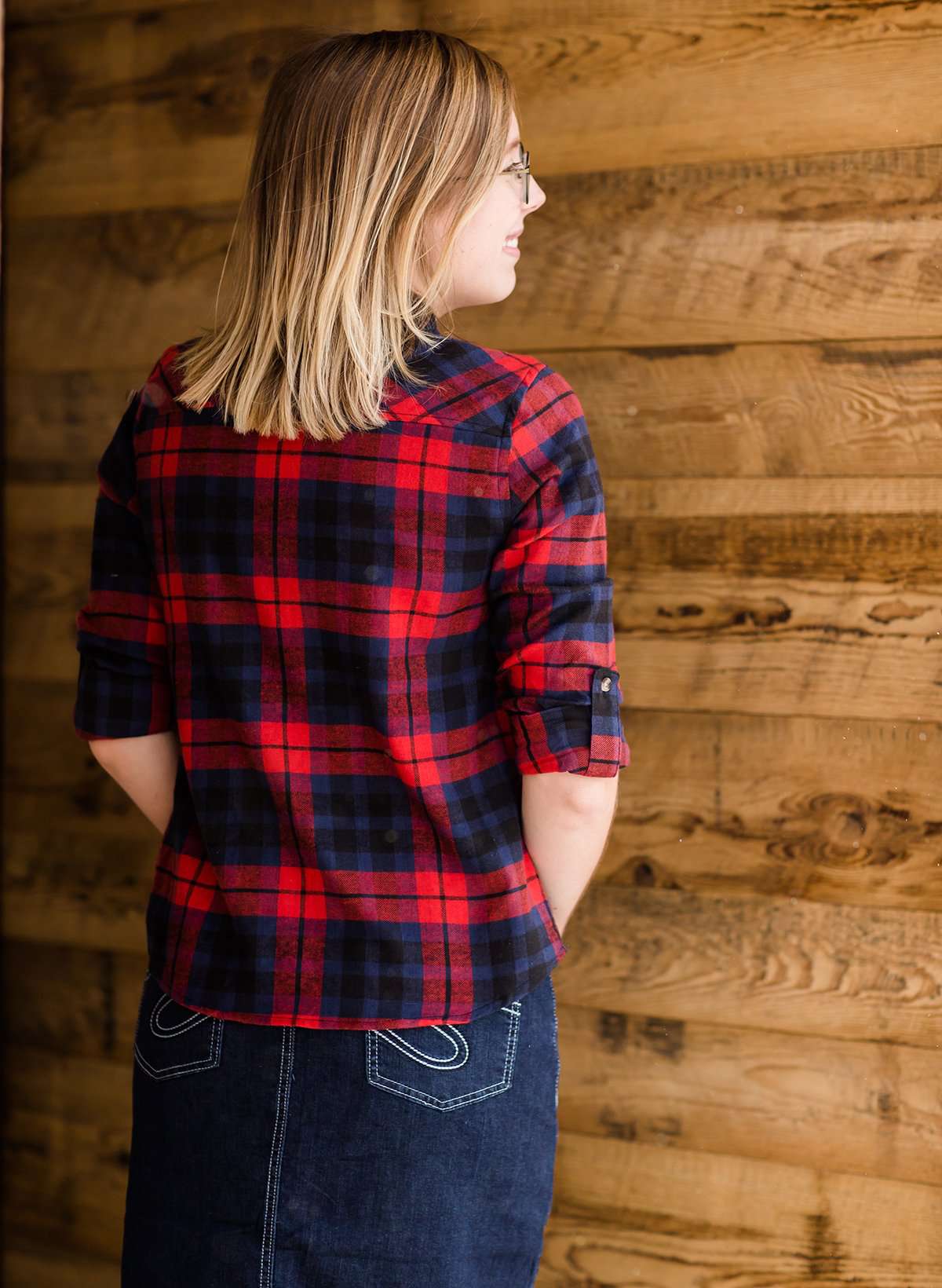 Woman wearing a cozy, checkered plaid top. This button up plaid shirt features roll tab sleeves and is black, navy and red. It is also paired with a below the knee jean skirt.