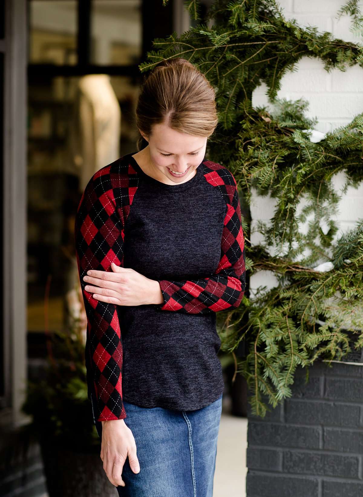 Woman wearing a dark charcoal raglan style top with diamond, argyle checks that are black and red. She is standing outside of Inherit Clothing Company.