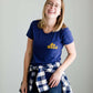 Young woman wearing a blue graphic tee with yellow words that says "Do Good." It is also paired with a below the knee denim skirt and a plaid button up top.
