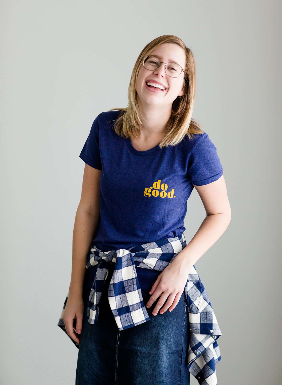 Young woman wearing a blue graphic tee with yellow words that says "Do Good." It is also paired with a below the knee denim skirt and a plaid button up top.