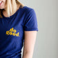 Young woman wearing a blue graphic tee with yellow words that says "Do Good." It is also paired with a below the knee denim skirt and a plaid button up top.