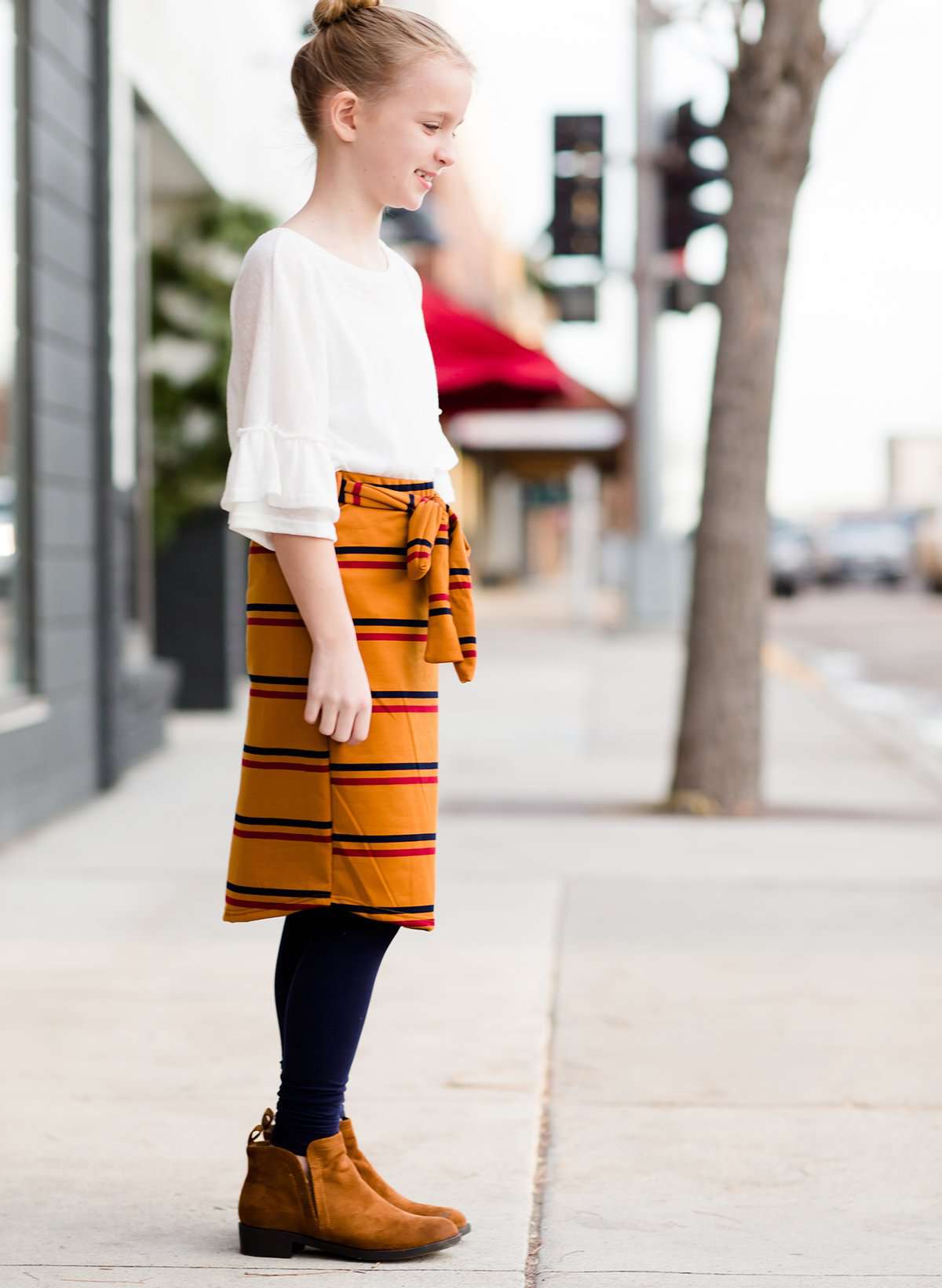 Young girl wearing an ivory, oversized modest top with ruffle sleeve accents. This top is also paired with a mustard, striped below the knee skirt and short boots.